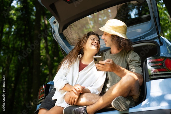Fototapeta Happy young couple sitting in open car trunk while travelling. Summer vacation and travel concept