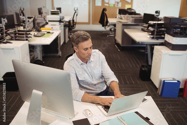 Obraz Middle-aged man in business casual attire typing on laptop at desk in open-plan office with monitor
