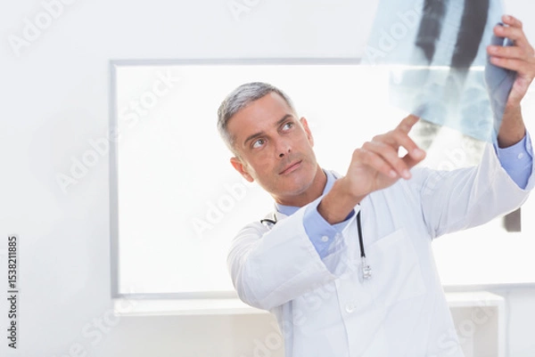 Obraz Male doctor in lab coat reading X-ray film on lightbox in clinic with stethoscope, copy space
