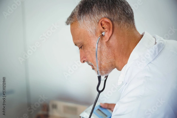 Obraz Medical professional listening with stethoscope while holding monitor against chest in clinic