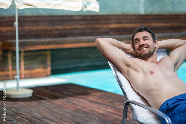 Obraz male wearing blue swim trunks reclining on pool deck lounge chair under umbrella, smiling relaxing