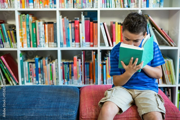 Fototapeta School-age boy sitting on blue and red bench reading green hardcover book in library, copy space