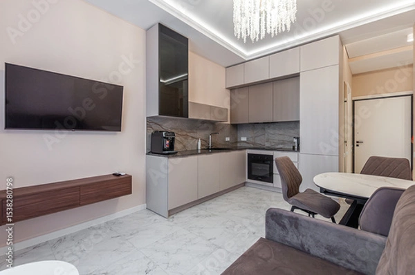 Fototapeta Living room with kitchen in small studio apartment with brown sofa, TV, nightstand, table with chairs, built-in refrigerator and cabinets.