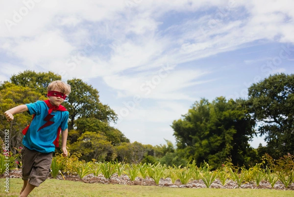 Fototapeta Boy wearing blue T-shirt showing red lightning bolt, red cape, eye mask running across grassy lawn
