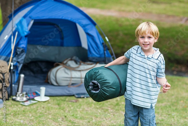 Obraz Boy carrying rolled sleeping bag while standing on grassy campsite by blue dome tent, copy space