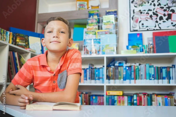 Fototapeta Boy reading open book at white table in school library reading corner with colorful books behind
