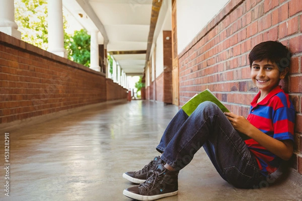 Fototapeta Hispanic boy reading green book while sitting on school corridor floor by brick wall, copy space