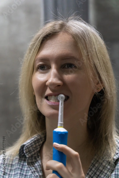 Obraz Young woman brushing her teeth with a modern electric toothbrush, maintaining good oral hygiene as part of her daily routine in a domestic bathroom setting. Vertical photo
