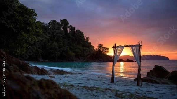 Fototapeta serene beach showcases a decorative arch draped with fabric as the sun sets over calm waters. Lush trees frame the background, creating a peaceful atmosphere in this tropical paradise