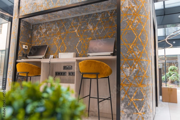 Obraz Stylish working area featuring a desk with two computers, mustard yellow bar stools, geometric wall design, natural light, and a courtyard view.