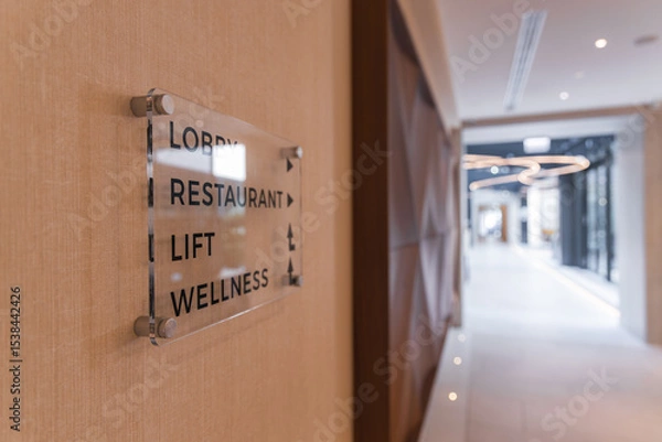Obraz Elegant hotel hallway featuring a directional sign, wooden paneling, circular light fixtures, and natural light from large glass windows.