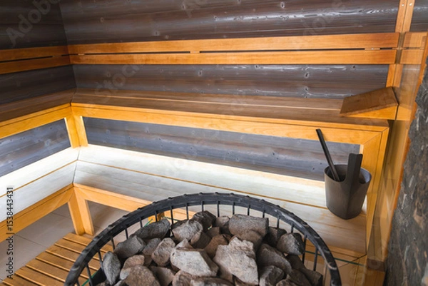 Obraz Luxury sauna featuring light wood benches, dark wood paneling, and a central heating element with stones, bucket, and ladle in a sleek design.
