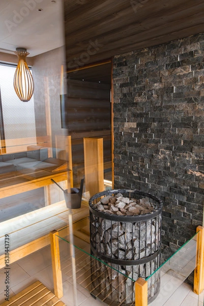 Obraz Modern sauna interior featuring sleek wooden benches, a stone wall, a central heating element with stones, and a warm wooden light fixture.