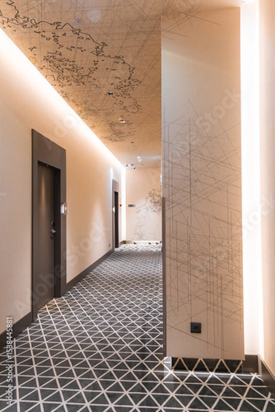 Obraz Elegant hotel hallway featuring a gray and white geometric carpet, map like ceiling artwork, recessed lighting, and dark colored doors.