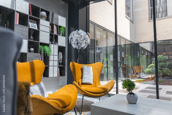 Obraz Luxurious hotel interior featuring mustard yellow armchairs, a sculptural lamp, sleek shelving, and large windows overlooking a serene courtyard.