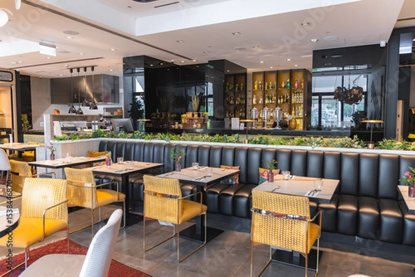 Obraz Elegant restaurant interior with yellow chairs, black banquette, floral decor, illuminated bar, open kitchen, greenery, and large windows with city views.