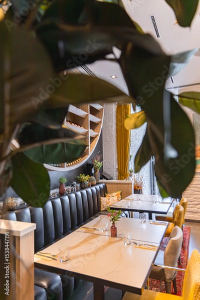 Obraz Dining area features marble topped tables, cushioned chairs, yellow curtains, a white brick wall, and a curved shelving unit with plants and decor.