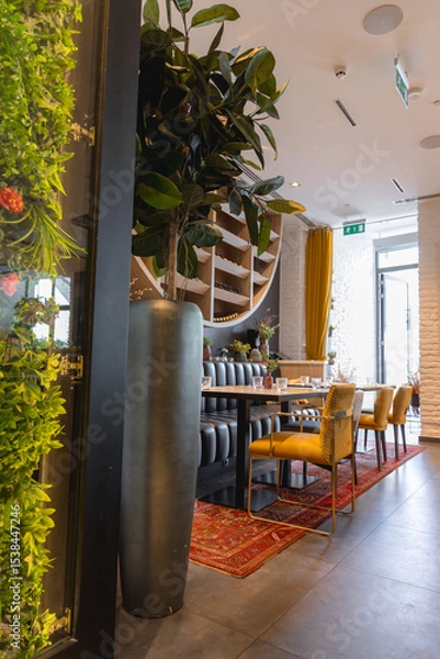 Obraz Luxurious dining space featuring a long table, mustard yellow chairs, tufted bench, red patterned rug, green wall, and natural light from large windows.