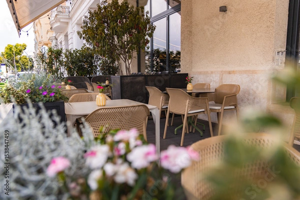 Obraz Outdoor seating area with beige wicker chairs, tables, potted plants, and flowers on a wooden deck. Beige stucco wall and glass windows frame the space.