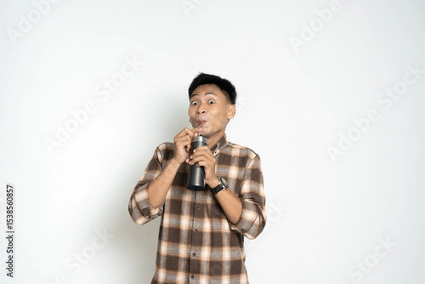 Fototapeta cheerful Asian man drinks from a bottle with a fun expression, wearing a casual plaid shirt, in front of a plain white background.