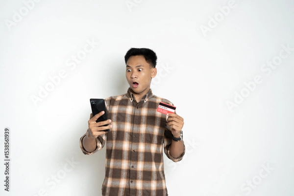 Fototapeta A man with a surprised expression holds a credit card and smartphone, showcasing emotion and modern lifestyle.