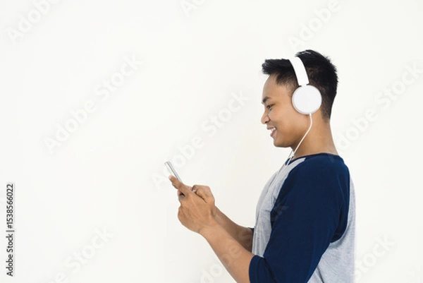 Fototapeta A young man smiles while using his smartphone and wearing white headphones, highlighting modern digital leisure, listening to music, and online connectivity.