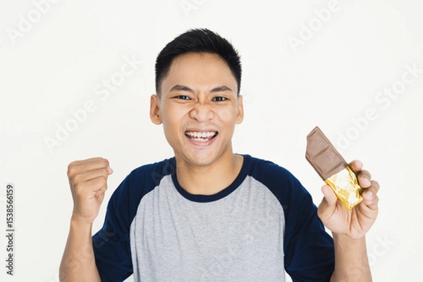 Fototapeta A joyful young man cheerfully holds a chocolate bar, showing excitement and enthusiasm.