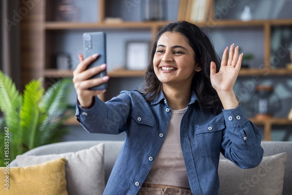 Fototapeta Smiling young Indian woman sitting on sofa at home, talking on video call and greeting on mobile phone camera