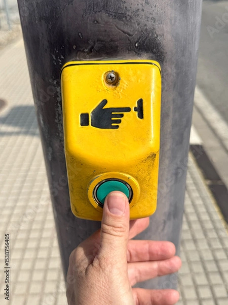 Fototapeta A hand pressing a yellow pedestrian crossing button on a pole. The button features a hand symbol and is located on a sidewalk.