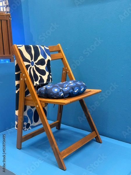 Fototapeta A wooden folding chair with a blue and white floral cushion. The chair is placed against a solid blue background, showcasing a modern design.