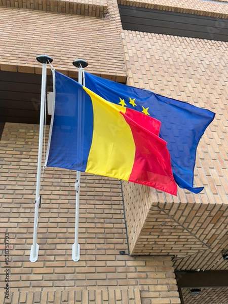 Fototapeta Flags of Romania and the European Union waving in the wind against a brick building background. Bright colors and clear design are visible.