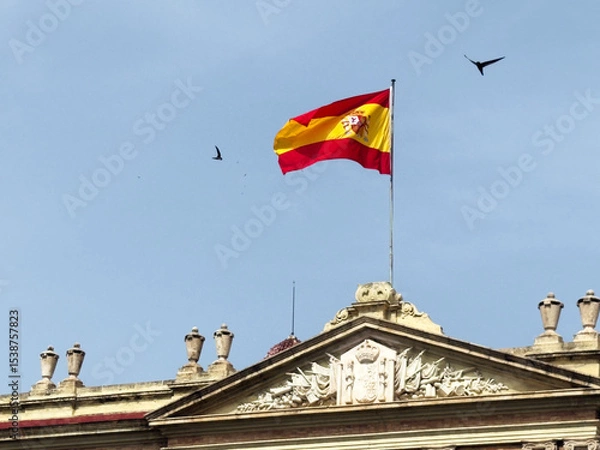 Fototapeta A Spanish flag waves atop a historic building under a clear blue sky. Birds fly in the background, adding to the serene atmosphere.