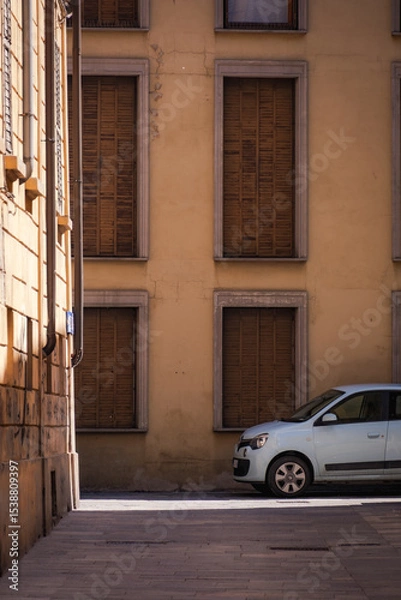 Obraz Reggio Emilia, Italy, 01.04.2025. Small light blue car parked near aged building facade with brown closed shutters