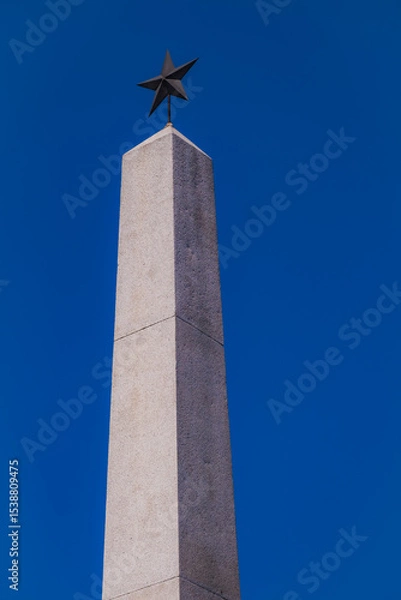 Obraz Reggio Emilia, Italy, 01.04.2025.Tall stone obelisk monument with metal star tip against deep blue sky, minimal composition