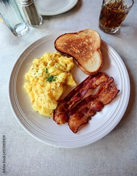 Fototapeta Delicious Breakfast Plate with Scrambled Eggs, Crispy Bacon, Toasted Bread, and a Drink on a Table in a Restaurant Setting