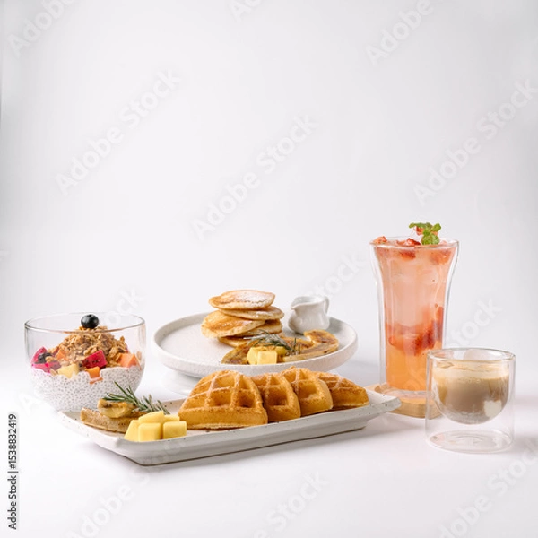 Fototapeta A stylish breakfast spread featuring waffles with butter, pancakes, chia pudding with berries, iced tea with mint, and a glass of coffee, set on a white background.