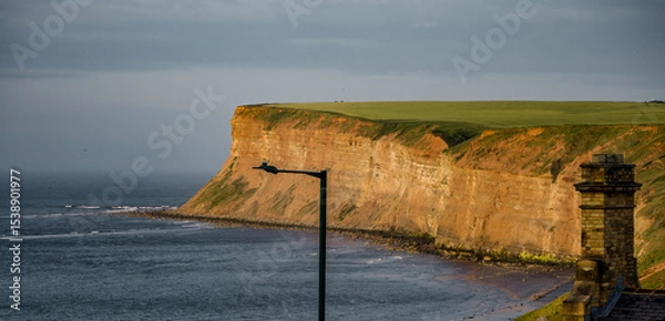Fototapeta Dramatic sunset light illuminating the cliffs at Saltburn by the Sea, UK, with a street lamp and chimney adding interest to the foreground