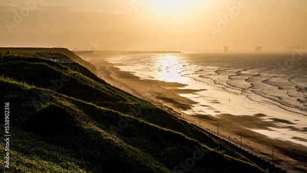 Fototapeta Breathtaking view of Saltburn beach during a golden sunset, with offshore wind turbines and grassy cliffs creating a serene coastal landscape