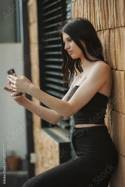 Obraz A young woman with long dark hair, dressed in a black crop top and pants, is sitting against a textured wall while focused on her smartphone