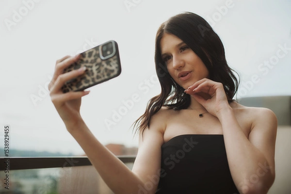 Obraz A woman with long dark hair enjoys taking a selfie on a balcony. She is dressed in a stylish outfit and poses playfully with a city skyline in the background
