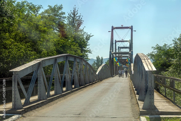 Fototapeta Historic Solotvyno Bridge connecting Ukraine and Romania, a symbol of cross-border connection, adorned with flags under a clear summer sky.
