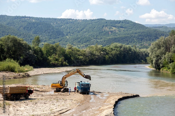 Fototapeta An excavator and dump trucks extract gravel from a riverbed, with lush green mountains and forests in the background on a sunny day.