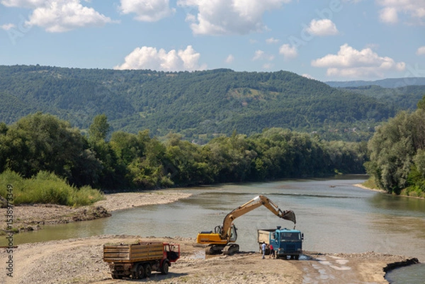 Fototapeta Industrial activity along a river bank: an excavator loads material into trucks, set against a backdrop of rolling forested hills