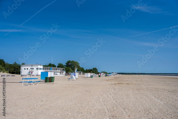 Obraz A serene beach in Parnu, Estonia, featuring a white building, lifeguard stands, and greenery under a clear blue sky. The coastline stretches into the distance.