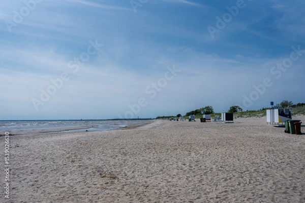 Obraz A tranquil sandy beach in Parnu, Estonia, with small structures near grassy dunes, a calm shoreline, and a faint horizon where sea meets sky.