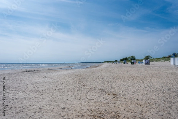 Obraz A serene sandy beach in Parnu, Estonia, features calm waters, a clear blue sky, scattered beach huts, sparse greenery, and low dunes in the background.