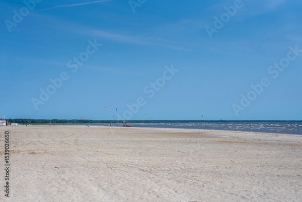 Obraz A tranquil beach in Parnu, Estonia, featuring calm waters, a clear blue sky, colorful kites in the distance, and a small structure on the left.