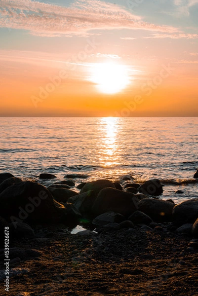 Obraz A serene sunset over the Baltic Sea in Parnu, Estonia, with warm orange hues, calm water reflections, a rocky shoreline, and wispy clouds in the sky.