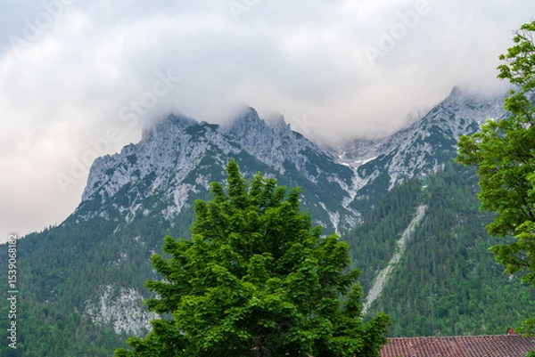 Fototapeta Zugspitze rises majestically through a veil of drifting clouds, its rugged, snow-dusted peaks cutting through the moody sky—Germany’s highest mountain cloaked in dramatic alpine mystery.