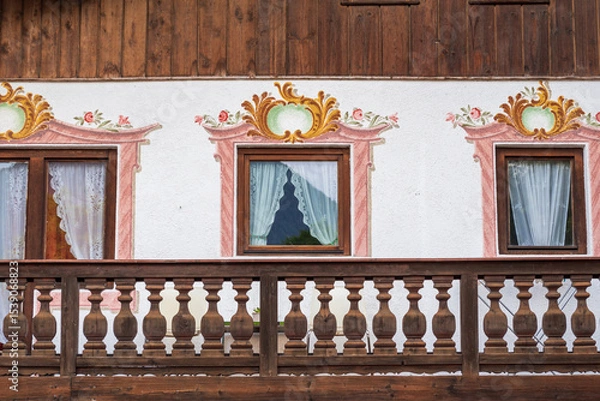 Fototapeta Charming alpine buildings in Mittenwald feature colorful Lüftlmalerei (frescoes), wooden balconies, and flower-filled window boxes, all set against a dramatic mountain backdrop.